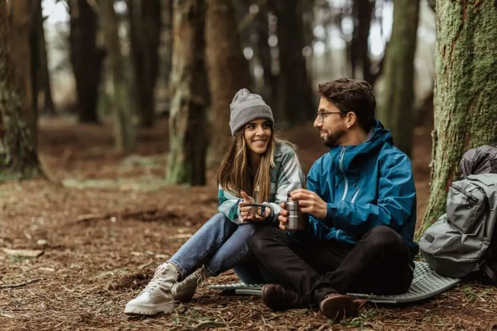 Glad young european guy and woman tourists in jackets with backpack rest in forest camping, enjoy